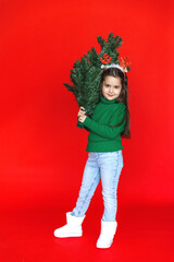 Beautiful cute little child girl in green warm sweater and reindeer horns holding little Christmas tree against red background. Christmas atmosphere, good overjoyed mood. High quality photo