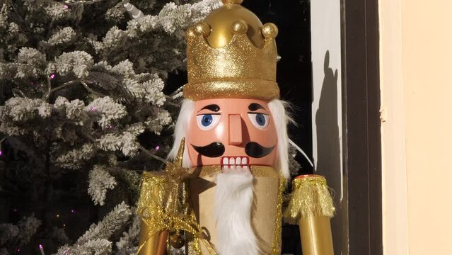 The Nutcracker big size doll dressed in a golden costume and crown, standing at the storefront window by the New Year tree, while people are passing by in the street.