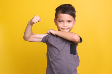 Funny Asian boy in casual gray t-shirt pulling up sleeve, showing muscle isolated at yellow...