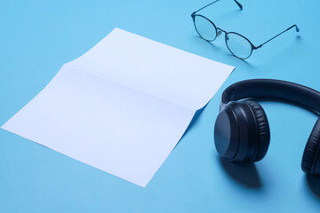 Headphones, glasses and blank white mockup paper on blue background