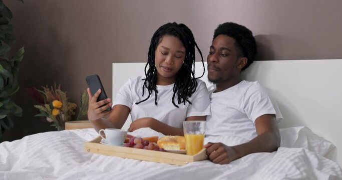 Lovely vute african amercan couple dark skinned girlfriend with boyfriend taking selfie picture having breakfast in bed. Happy relationship. Just married couple.