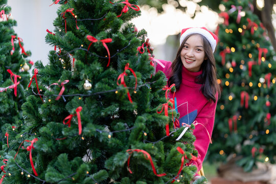 Young Asian woman in Santa hat smiling and holding Christmas gifts smile happily While decorating the Christmas tree at home, the idea of ​​celebrating New Year's Day