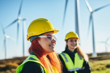 Two female engineers in hard hats and reflective vests observing wind turbines at sunset, representing renewable energy and teamwork