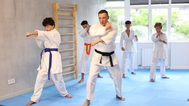 Teenagers in kimonos practicing effective karate techniques in group workout with their trainer in foreground at modern training room