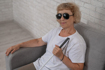 An elderly blind woman wearing sunglasses and with a tactile cane sits on the sofa. 