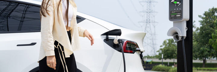 Young woman recharge EV car battery at charging station connected to power grid tower electrical...