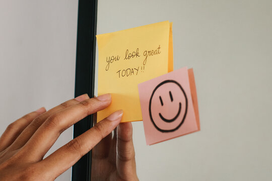 Woman putting a sticky note with phrase You Look Great Today! on the mirror.