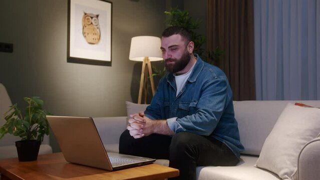 Handsome young adult bearded man at home at night sitting on sofa while having video call conversation through using laptop, share news, talk to family living abroad, enjoy webchat virtual meeting