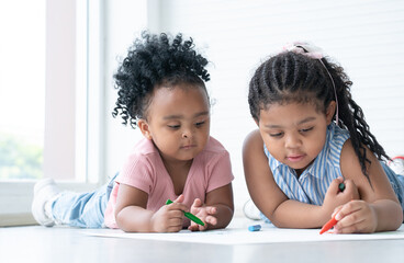 Fototapeta premium African cute little kid girl teaching her sister drawing on paper while lying on the floor. Adorable children have fun drawing with colorful crayons at home. Education, family lifestyles, creativity