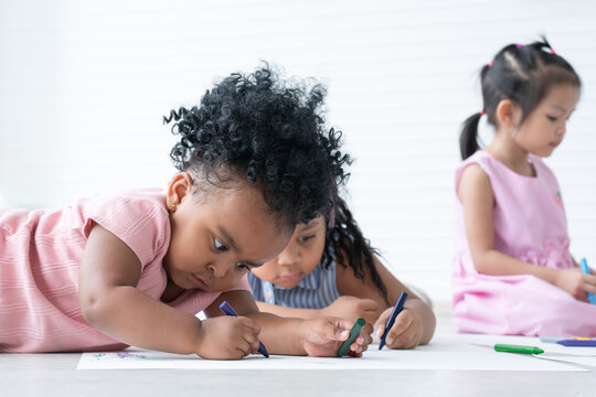 African cute little kid girl concentrate with drawing on paper while lying on the floor with friend. Adorable children drawing with colorful crayons at school. Selective focus on hand holding crayon