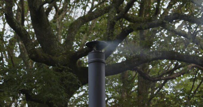 mid shot of a metal flue chimney with light smoke, large tree behind