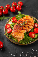 Grilled chicken breast with arugula and tomatoes on a plate and black table vertical photo