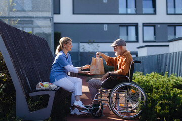 Nurse eating lunch outdoors with senior man in wheelchair. Female caregiver and elderly man enjoying a warm autumn day in nursing home, having picnic. - obrazy, fototapety, plakaty