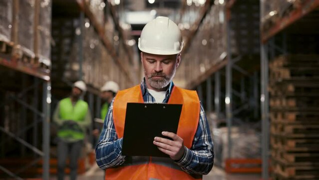 Handsome Caucasian Auditor Wearing Hard Hat Looking around Warehouse and Writting Down Information. Rows of Storage Racks are Seen. Logistic Business: Shipping and Delivery Service