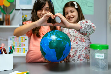 Portrait of two happy female students showing a handmade globe world earth while making heart shape...