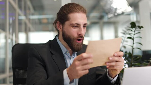 Positive caucasian professional man celebrating business success while holding envelope with good news inside. Emotional male feeling happy because signing employment contract at personal workplace.