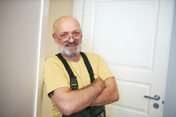 Happy senior bald and bearded caucasian repairman in glasses standing in doorway with crossed...