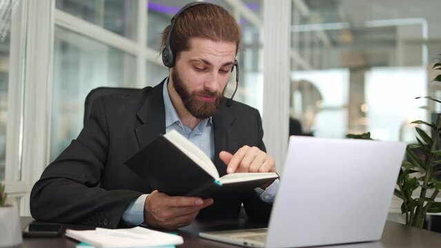 Handsome bearded male worker dressed in formal suit holding own notes and explaining them during video call with colleagues at workplace. Attentive businessman having online conversation indoors.