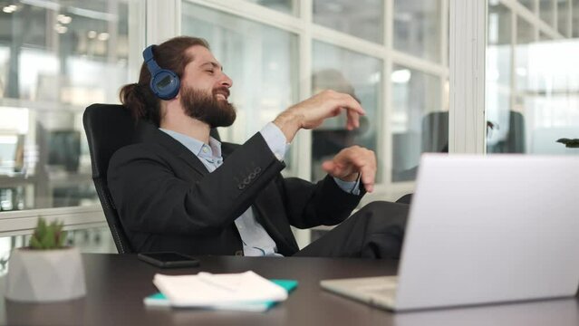 Smiling caucasian businessman wearing wireless headset imagining himself as orchestra conductor while waving virtual baton. Active male melomaniac listening classic music and putting feet on table.