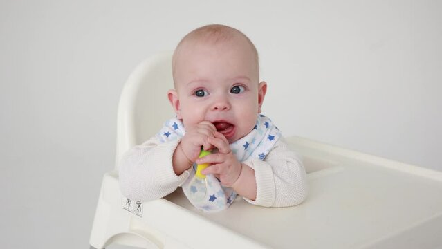 4k, cute baby girl sitting on a feeding chair nibbling a silicone baby spoon looking at the camera, the concept of introducing complementary foods for babies