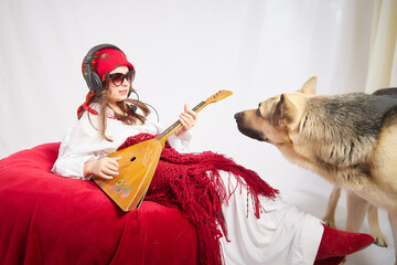 A girl in red scarf, white shirt, with shawl and balalaika in black sunglasses with a shepherd dog....
