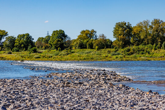 Abundant gravel and stones in Maas river, flowing water, alluvial terrain, Belgian countryside with trees in background, sunny day, blue sky in Maasvallei nature reserve in Meers, Elsloo, Netherlands