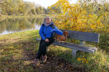 Fototapeta premium Senior hiker taking a break, sitting on wooden bench with her dog, river surrounded by autumn trees in background, Belgian nature reserve De Wissen Maasvallei, sunny day in Dilsen-Stokkem, Belgium