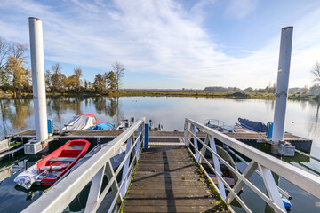 Bridge towards pier on lake with anchored boats, flat field against blue sky in background,...