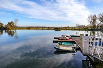 Lake with wooden pier with anchored boats, flat field against blue sky in background, reflection in...
