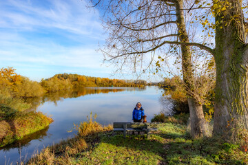 River surrounded by autumn trees against blue sky in background, female hiker standing with her dog next to a bench, Belgian nature reserve De Wissen Maasvallei, sunny day in Dilsen-Stokkem, Belgium