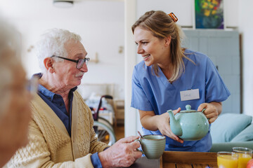 Happy healthcare worker holding teapot and talking to senior man at table