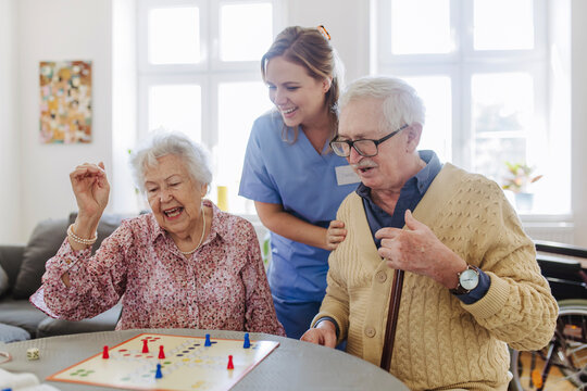 Happy senior couple playing ludo game with healthcare worker standing at home