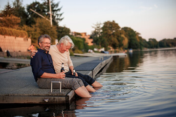 Happy senior friends sitting on jetty with beer bottles near lake
