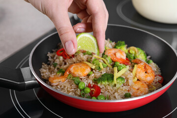 Woman squeezing lime into rice with shrimps and vegetables on induction stove, closeup