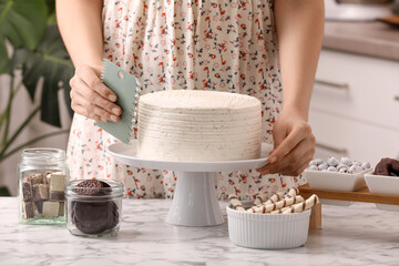 Woman using scraper to decorate cake at white marble table in kitchen, closeup - obrazy, fototapety, plakaty