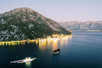 Illuminated Church of Our Lady on the Rocks on the island against the backdrop of illuminated Perast. Montenegro. Drone