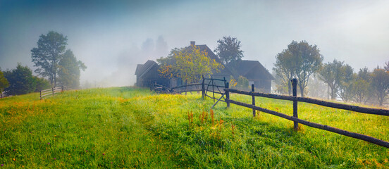 Sunlight breaks through the fog onto a green meadow in a mountain village. Panoramic summer view of...