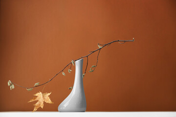Vase with tree branch and autumn leaf on white table against brown background. Space for text