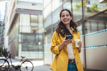 Woman texting and drinking coffee outdoors. Young smiling brunette woman holding a smartphone and a...