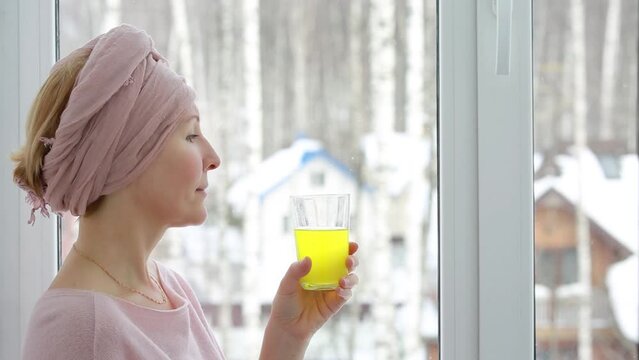 A woman in a turban drinks a soluble vitamin drink of yellow color against the background of winter outside the window.