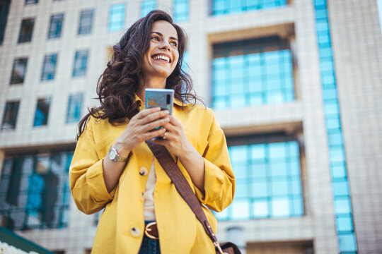 Beautiful happy young woman messaging on smartphone on the background of a city street on a sunny day. Business woman is looking away and smiling. 