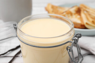 Tasty condensed milk in jar and crepes on white table, closeup