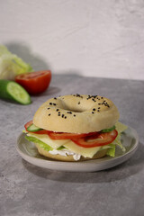 Delicious bagel with vegan cheese and fresh vegetables on a plate, grey table