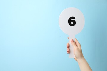 Woman holding auction paddle with number 6 on light blue background, closeup. Space for text