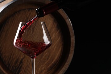 Pouring red wine from bottle into glass near wooden barrel against black background. Space for text