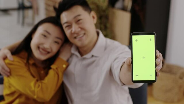 Blurred asian couple bonding at home and showing smartphone with green chroma key screen and tracking marks, empty space