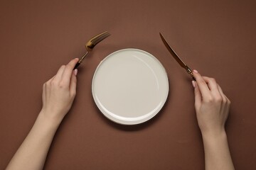Woman holding fork and knife near empty plate at brown table, top view