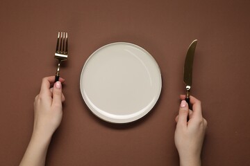Woman holding fork and knife near empty plate at brown table, top view