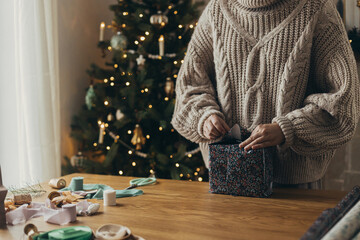Woman in cozy sweater wrapping stylish christmas gift in festive wrapping paper with ribbons,...
