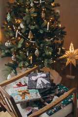 Merry Christmas! Stylish christmas gifts wrapped in festive paper with bows, vintage ornament and toy on modern chair on background of christmas tree lights. Atmospheric winter holidays
